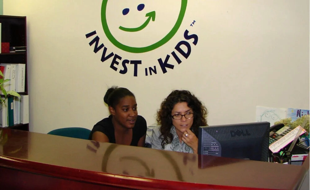 Maxine Paynter and a colleague looking at a computer screen with the Invest in Kids logo on the wall behind them.